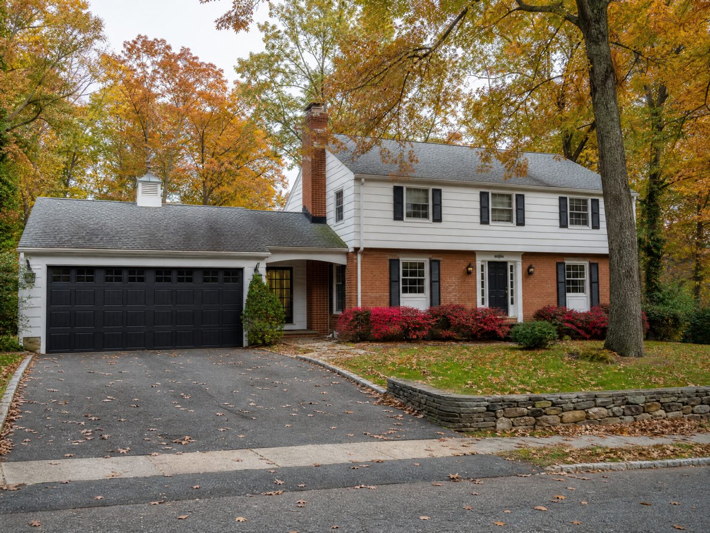 A charcoal carriage-style garage door on a brick colonial with large oak trees and fallen leaves on the driveway