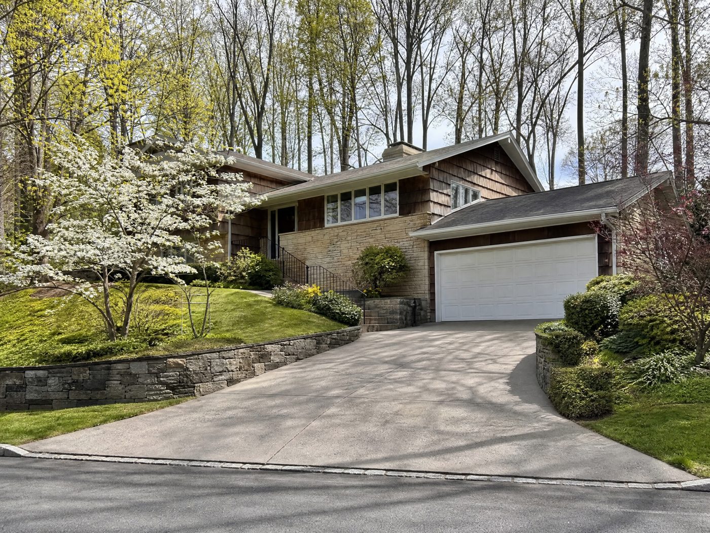 A white raised-panel garage door on a stone and cedar split-level with a curved sloped driveway and flowering dogwood